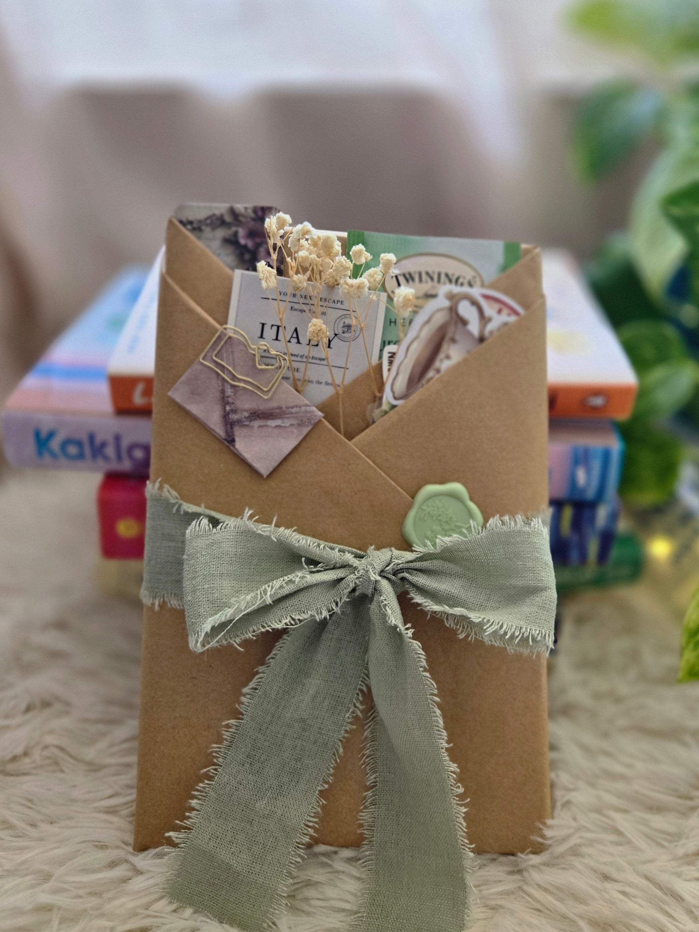 A wrapped gift box in brown kraft paper with a sage green ribbon bow, decorated with small vintage-style paper tags, resting on a white textured rug with a blurred indoor background.